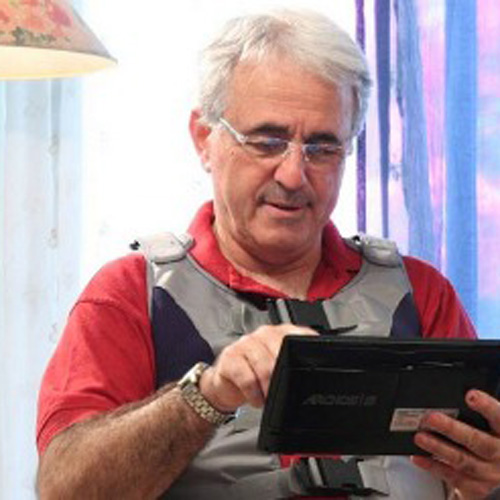 An older man with glasses and a red shirt sits, using a digital device. He wears a light-colored vest with a strap. A lamp and sheer curtains are in the background, providing a cozy indoor atmosphere.