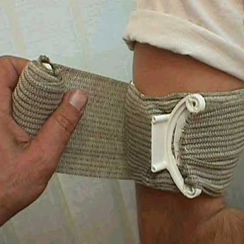 A person wraps a beige elastic bandage with a plastic fastener around their elbow for support. The bandage covers the joint area, and the persons thumb holds it in place while wrapping. The person is wearing a white shirt.