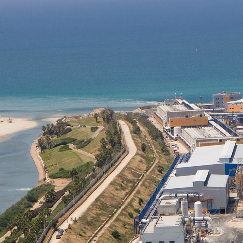 A coastal industrial facility with large buildings is situated along a shoreline. The complex is adjacent to a narrow strip of greenery and palm trees, with the ocean stretching out in the background. The scene is set under a clear blue sky.