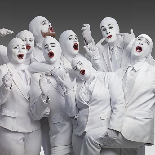 A group of performers dressed in all-white suits, wearing white face paint and red lipstick. They are striking expressive poses, showcasing a theatrical, mime-like style against a plain gray background.