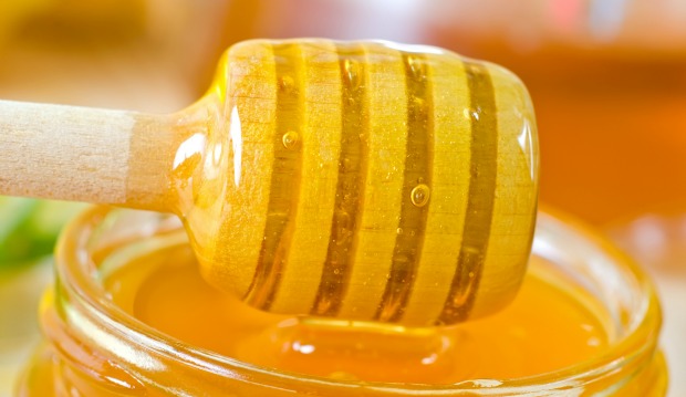 A close-up of a honey dipper coated with golden honey, resting above an open jar filled with honey. The background is softly blurred in warm tones.