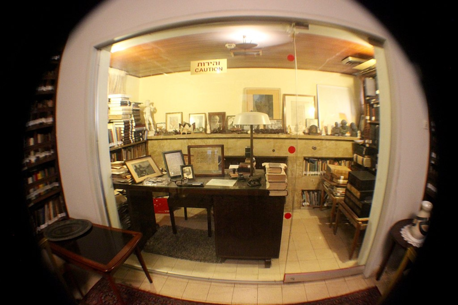 Gabi Davis took this photo inside the Ben-Gurion House in Tel Aviv. Photo via Wikimedia Commons A fisheye lens view of an office in Ben Gurion house filled with books, stacks of paper, lamps, and framed photographs. The room has a glass partition with a Caution sign in Hebrew, showcasing a cozy, cluttered workspace.