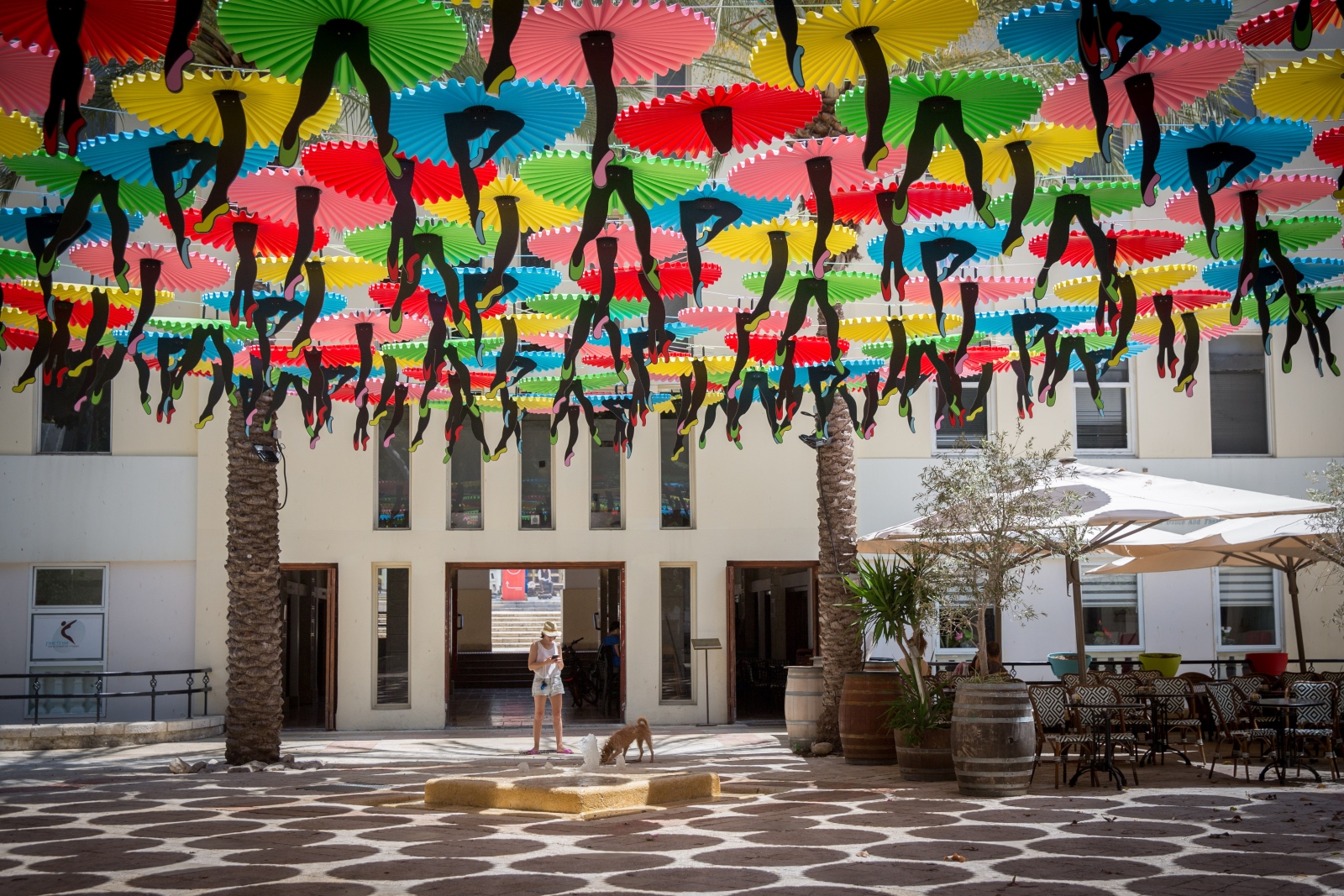 Colorful umbrellas with "dancing feet" hang at the plaza outside the Suzanne Dellal Center in the Tel Aviv neighborhood of Neveh Tzedek8. Photo by Miriam Alster/FLASH90 A courtyard in Tel Aviv with colorful umbrellas hanging overhead, creating shade. A person stands near a bronze dog statue on a tiled floor with circular patterns. Palm trees and outdoor seating with umbrellas surround the space.