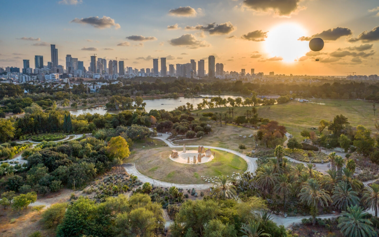 A view of the rock garden in Yarkon Park. Photo by Seth Aronstam, Shutterstock Aerial view of a Tel Aviv's skyline at sunset with Yarkon Park in the foreground. The park features green fields, circular paths, and clusters of trees. The city skyline and a partly cloudy sky with the sun setting are in the background.