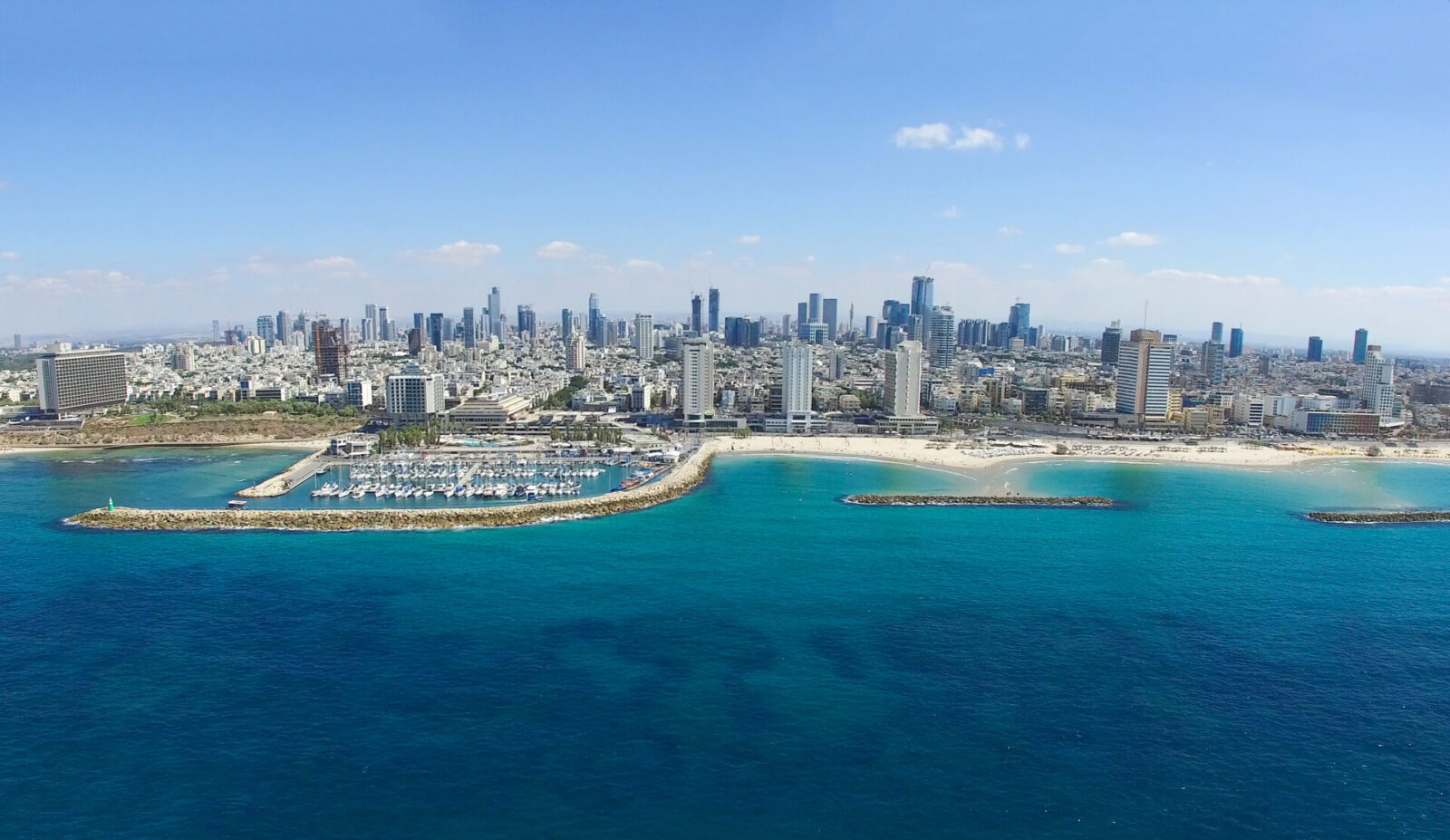 Aerial view of Tel Aviv's modern city skyline with tall buildings along a coastline. A marina is visible next to a sandy beach, bordered by clear turquoise water under a clear blue sky.