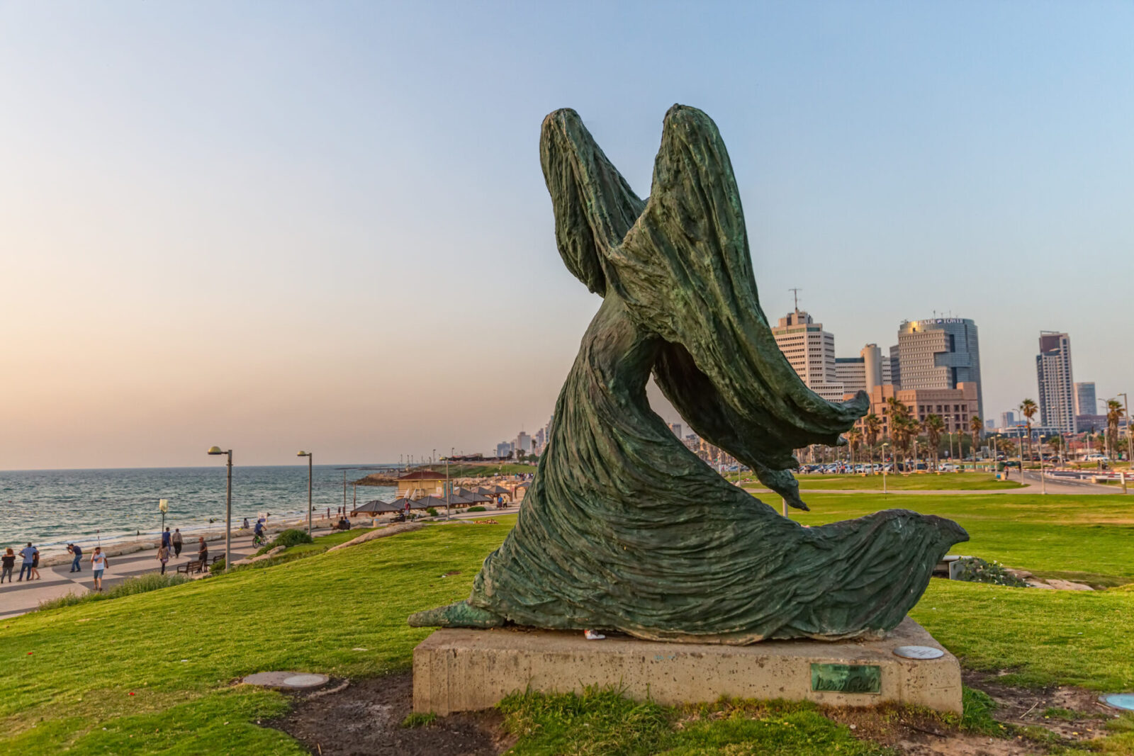 Woman against the wind sculpture at Charles Clore Park in Tel Aviv. Photo by OPIS Zagreb, Shutterstock Bronze statue of a woman in motion, draped in flowing clothing, set in a Charles Clore Park by the seain Tel Aviv. Skyscrapers line the horizon under a clear blue sky.