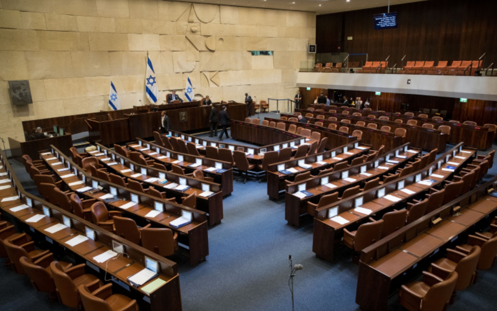 As well as rowdy politics, Israel’s parliament is home to fascinating artwork and architecture. Photo by Yonatan Sindel/Flash90 Empty legislative chamber within the Knesset with rows of brown seats and desks, featuring blue and white flags with the Star of David. Walls are beige stone, and a small gallery is visible. Large emblem is mounted on the wall.