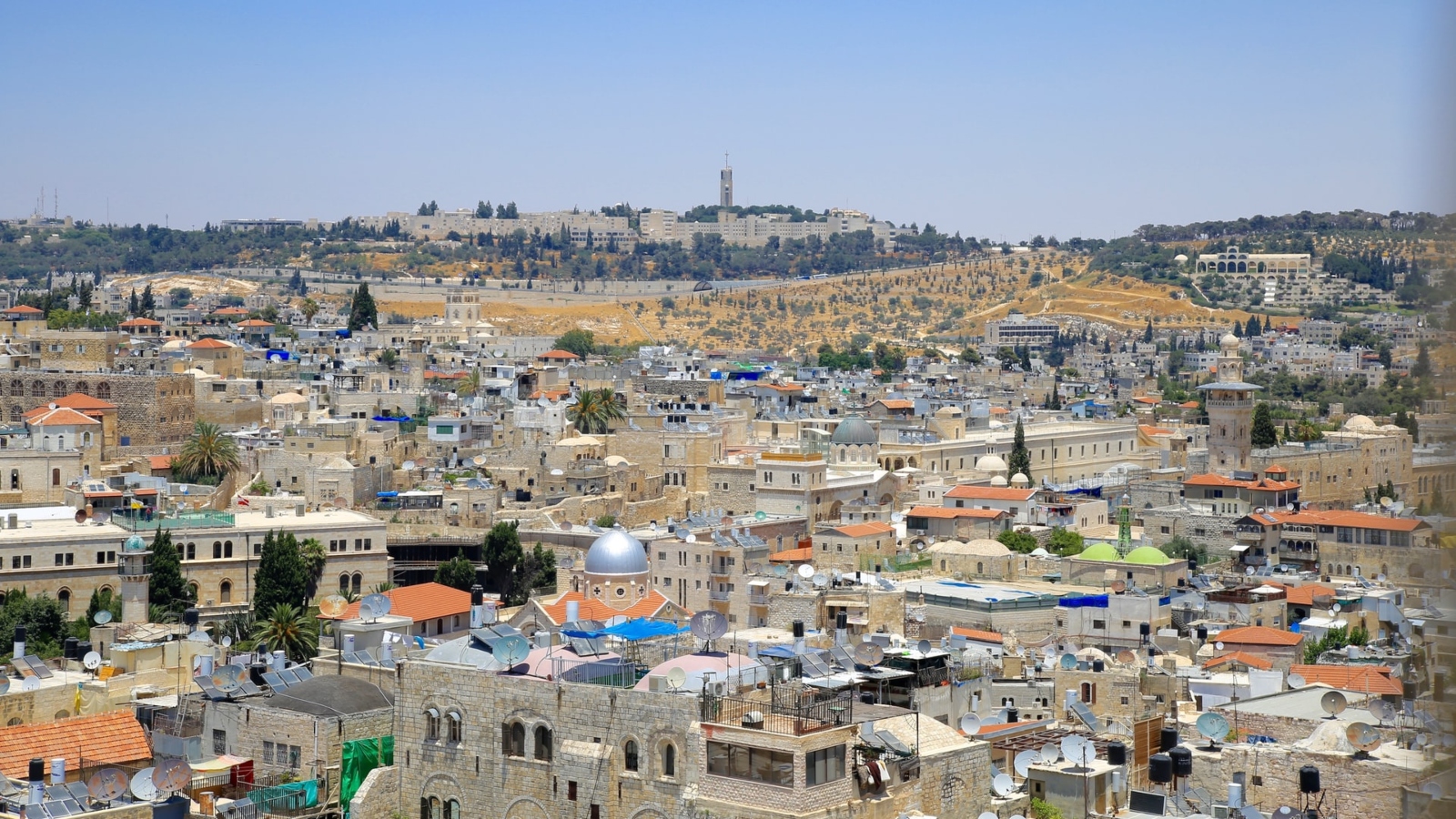 A panoramic view of a densely the built Middle Eastern city of Jerusalem, with a mix of modern and historical architecture. The scene includes domed rooftops, a hill with scattered buildings, and a clear blue sky above.