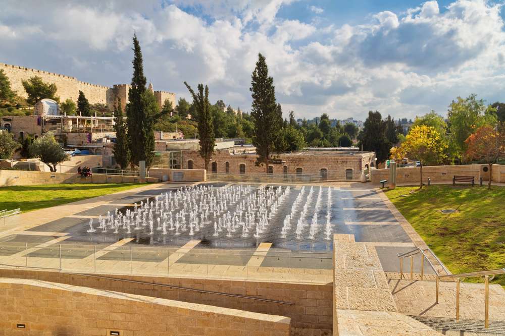 Teddy Park is one of the rare meeting places for all parts of Jerusalem society, particularly kids. Photo by DeltaOFF / Shutterstock.com A scenic view of Teddy Park with water fountains arranged in a grid. Trees surround the area, and stone walls are visible in the background under a partly cloudy sky. Benches and pathways are interspersed throughout the park.