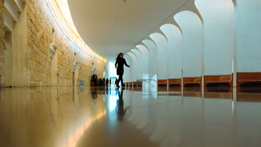 Israel’s Supreme Court is an architectural beauty that can be enjoyed via an excellent free tour. Photo by Shalom Rufeisen / Shutterstock A person walks through the spacious interior of the Supreme Court halls with a shiny, reflective floor. The left side features a stone wall with a curved design, while the right side has a white, arched structure. Soft, warm lighting illuminates the scene.