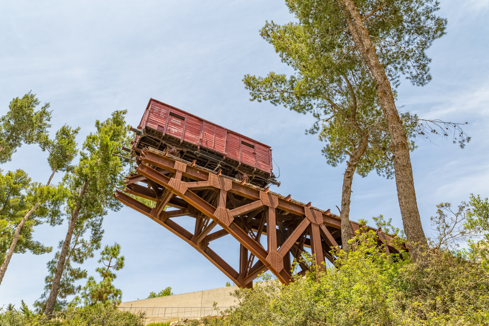 The Yad Vashem Holocaust Remembrance Center is full of remarkable exhibitions and includes this train used to transport Jews during the Holocaust. Photo by Opis Zagreb / Shutterstock A red train car perched on a rusted, elevated wooden trestle, surrounded by tall green trees against a blue sky. The structure sits outside of the Yad Vashem building, whose top is slightly visible towards the bottom of the photo, and is set amidst dense foliage.