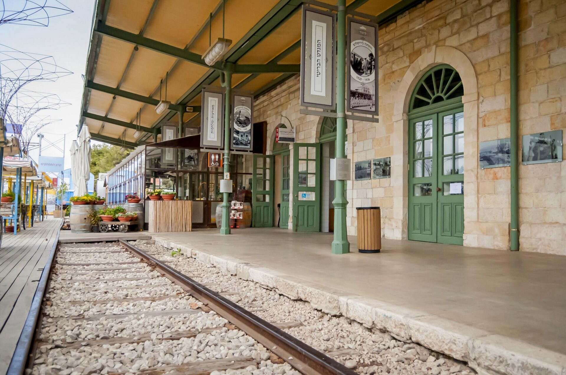 The First Station compound is lovely to visit night or day, and hosts a variety of free and interesting events. Photo by Roman Yanushevsky, via Shutterstock The vintage looking First Station railway station in Jerusalem, with a stone facade, green doors, and arched windows. The platform features old-style lamps and signs. Train tracks run alongside, and potted plants add color to the scene. The area appears quiet and serene.