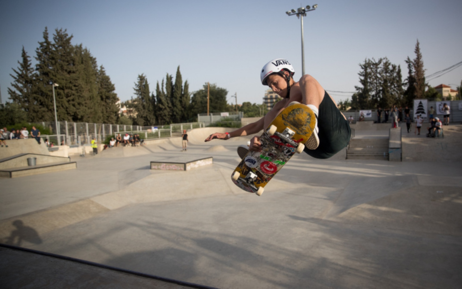 Jerusalem’s skate park is a hub of activity for local kids, teens and families. Photo by Yonatan Sindel/Flash90 A young boy skateboarding mid-air in an outdoor skatepark, wearing a helmet and holding the skateboard with one hand. The background features trees, lampposts, and other people in the park. Its a bright, clear day.