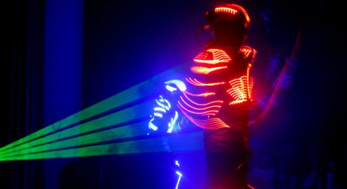 A performer in a suit with glowing red lights stands on stage, facing away, while green laser beams shoot out. Another performer in blue LED lights is visible in the background against a dark backdrop.