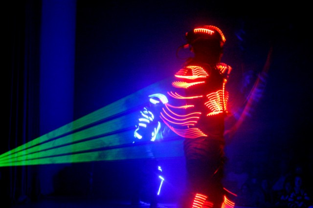 A person in a suit with glowing red lights stands on stage, facing away, while green laser beams shoot out. Another person in blue LED lights is visible in the background against a dark backdrop.