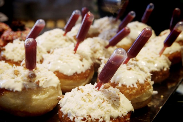 Rows of donuts topped with white icing and shredded coconut, each donut featuring a small plastic pipette filled with red liquid inserted into the center.