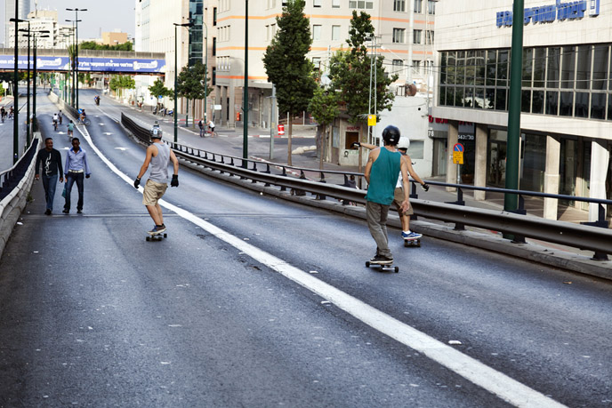 Three people ride skateboards down a wide, empty city street in Tel Aviv, while two pedestrians walk toward them. Tall buildings, trees, and streetlights line the road in the background.