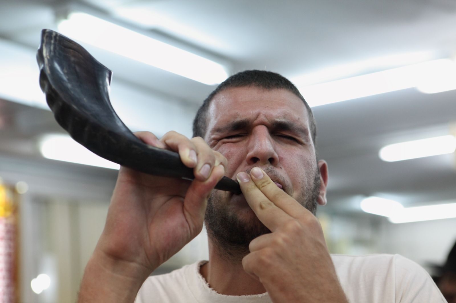 Blowing a shofar at the Rabbi Basri Yeshiva in Beersheva. Photo by Yaakov Naumi/FLASH90 A man with short hair and a beard blows a traditional shofar horn, eyes closed and fingers near his mouth, inside a brightly lit room.