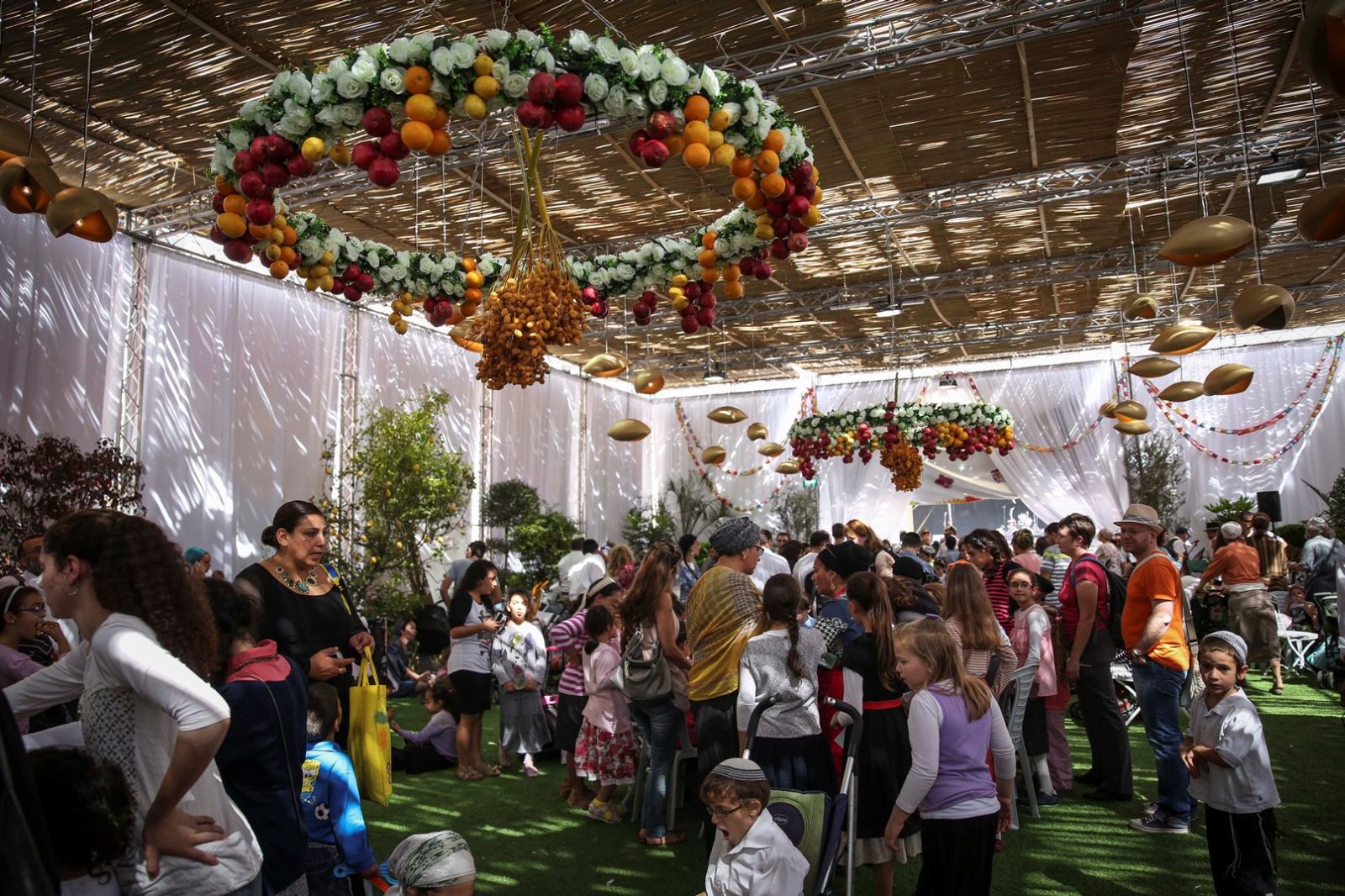 Visitors pack Jerusalem’s Safra Square sukkah every year. Photo by Flash90 A large group of people, including many children, gather under a decorated Sukkah with hanging fruit arrangements and white drapes. Sunlight filters through the roof, creating patterns on the crowd and artificial grass floor.