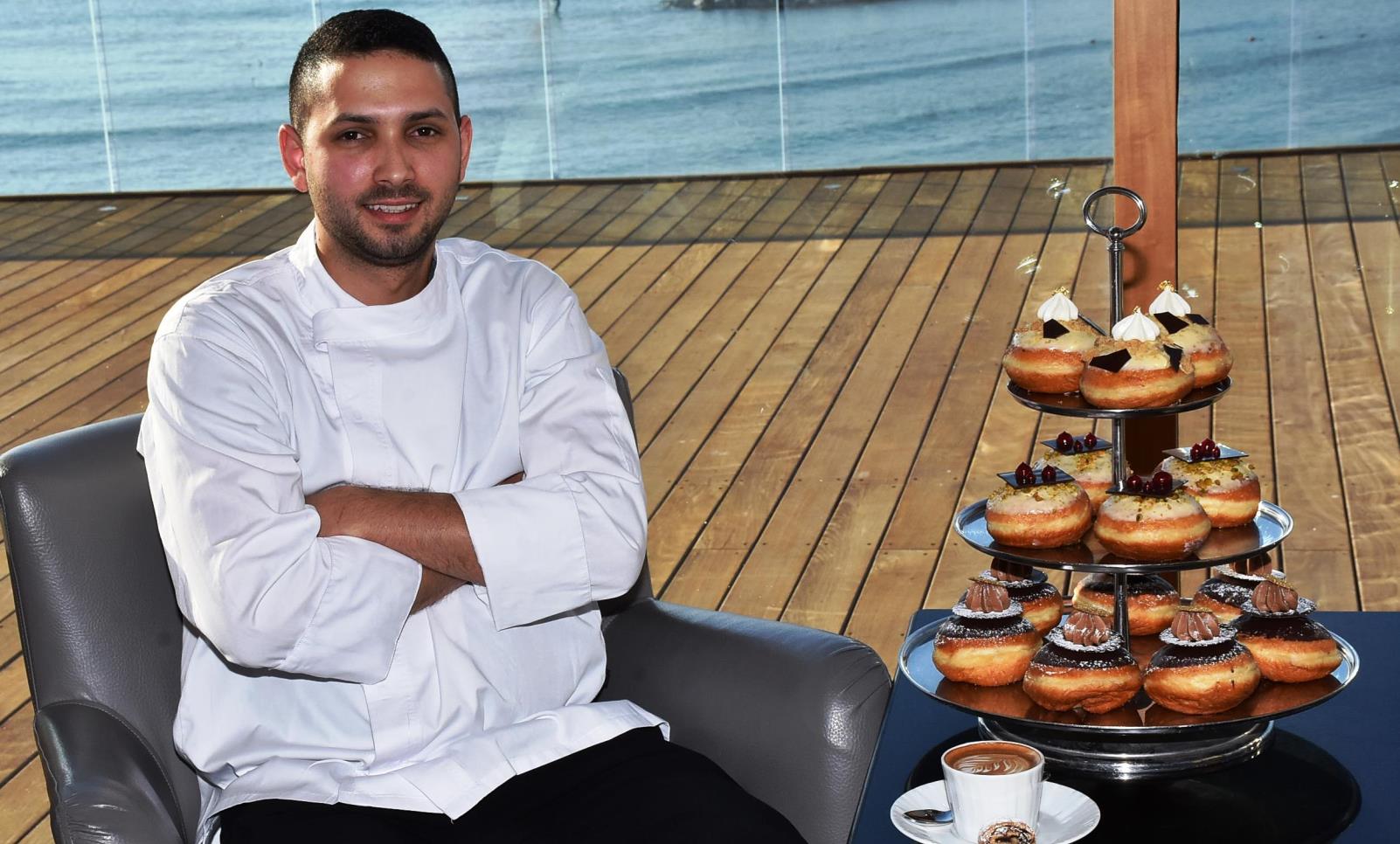 Tel Aviv pastry chef Idan Hadad, in a white uniform sits on a chair by a wooden deck overlooking water. Beside him is a tray stand filled with assorted sufganiyot and a cup of coffee on a small table.