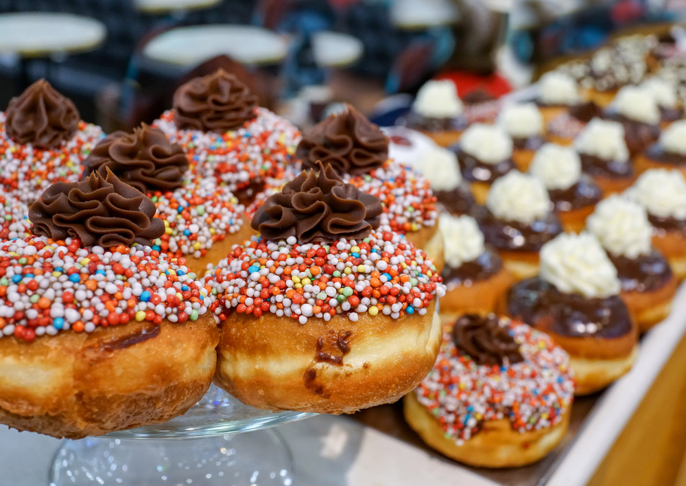 A close-up of donuts topped with colorful sprinkles and swirls of chocolate frosting, displayed on a glass tray. More donuts with various toppings are blurred in the background.