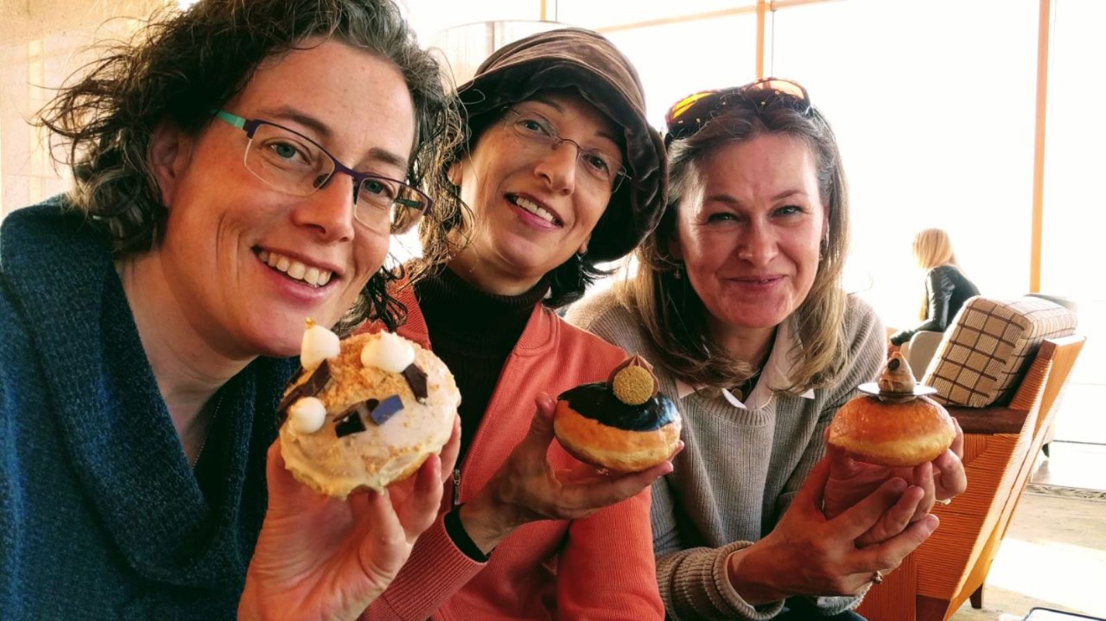 Three smiling women holding decorated Hanukkah donuts, sitting close together in a bright, indoor setting. Each woman is presenting her donut toward the camera, enjoying a casual, cheerful moment together.