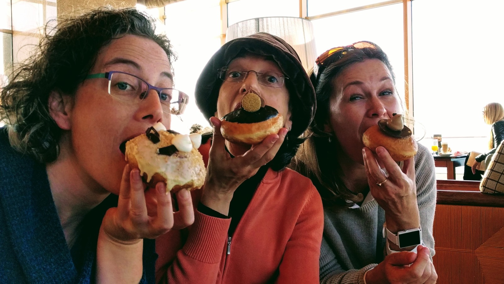 Three women smiling and holding decorated Hanukkah donuts up to their mouths as if about to take a bite. They appear to be having fun together in a bright indoor setting.