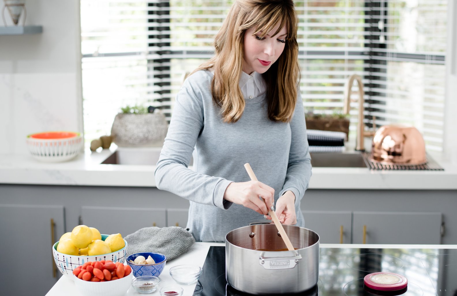 Chef Jamie Geller uses date honey in both sweet and savory dishes. Photo: courtesy A woman stands in a modern kitchen stirring food in a pot on the stove. Nearby are bowls of tomatoes, lemons, garlic, and a blue cloth on the white countertop under bright natural light.
