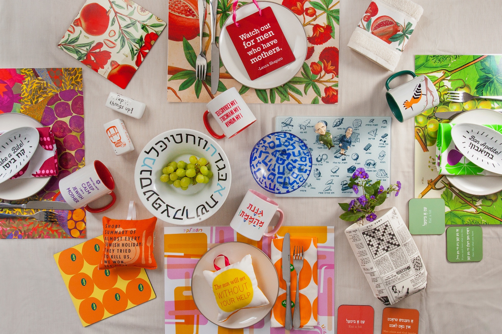 Barbara Shaw Gifts will quell your Israel homesickness. Photo: courtesy A colorful table set with vibrant placemats, plates, cups, napkins, and small gifts from Barbara Shaw designs, many featuring Hebrew text, fruit designs, and playful patterns. Grapes and flowers are also placed among the table settings.