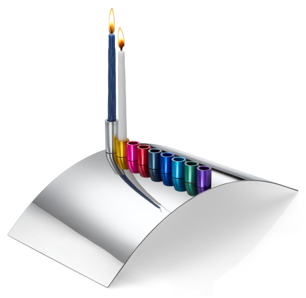 Building Bridges menorah by Laura Cowan Modern Judaica. Photo: courtesy A modern, curved silver menorah holds two lit candles—one blue and one white—with the remaining colorful candle holders empty, reflecting on the shiny surface.