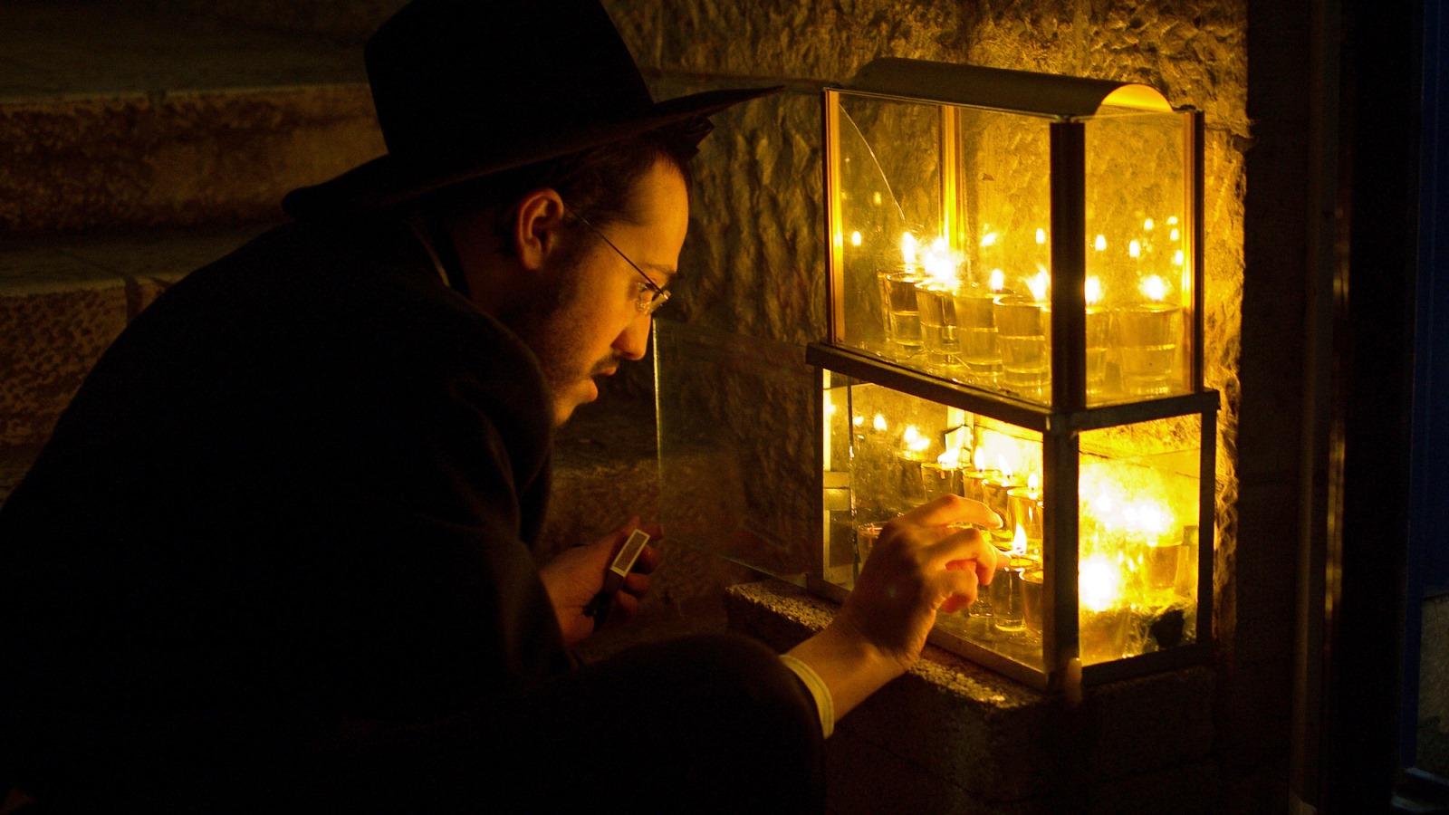 A man wearing a black hat and glasses is lighting candles of a menorah inside a glass case during a nighttime Hanukkah celebration. The candlelight illuminates their face and hand, creating a warm, intimate atmosphere.