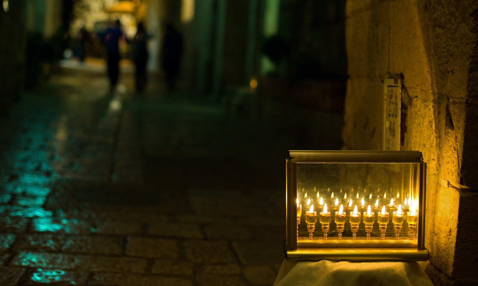 A menorah with lit candles is displayed in a glass case on a stone street at night, with a softly illuminated walkway and blurred figures in the background.
