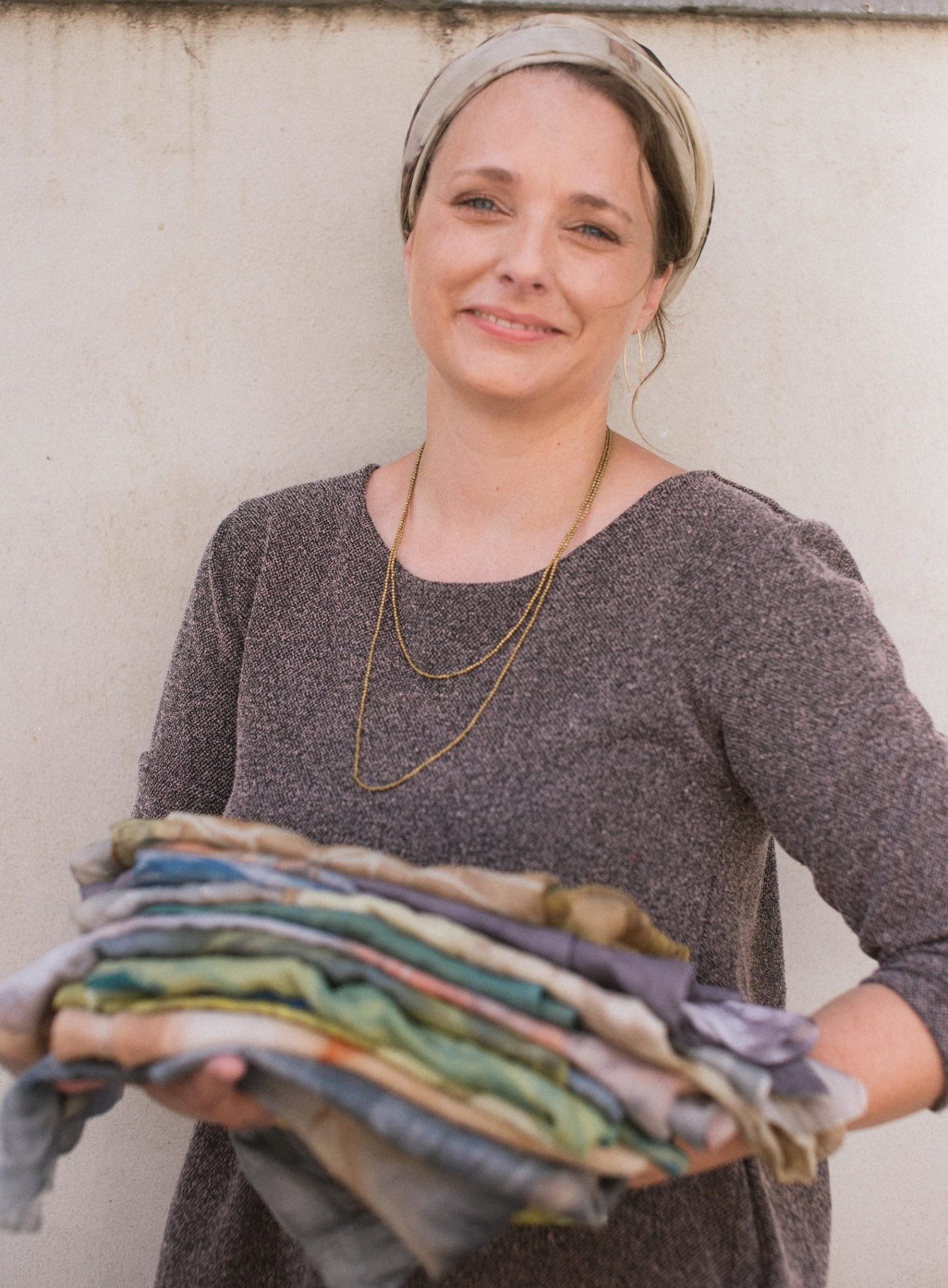Suzanne Dekel Botanical Prints. Photo by Becky Kestenbaum A woman with a headscarf and a brown dress smiles while holding a large stack of colorful folded fabrics in her hands, standing against a plain light-colored wall.