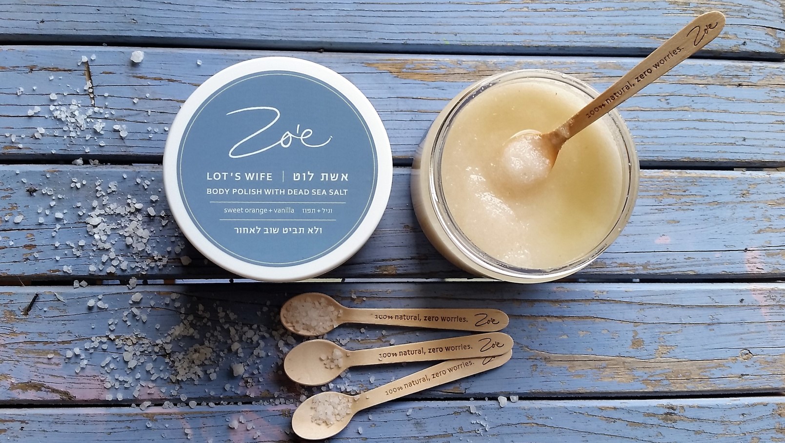 Lot’s Wife body polish is infused with orange and vanilla. Photo by Layla Levy/Zoe Apothecary An open jar of body polish with a wooden spoon, next to a closed lid labeled Zoe Lots Wife Body Polish with Dead Sea Salt, three smaller wooden spoons, and sea salt scattered on a blue wooden surface.