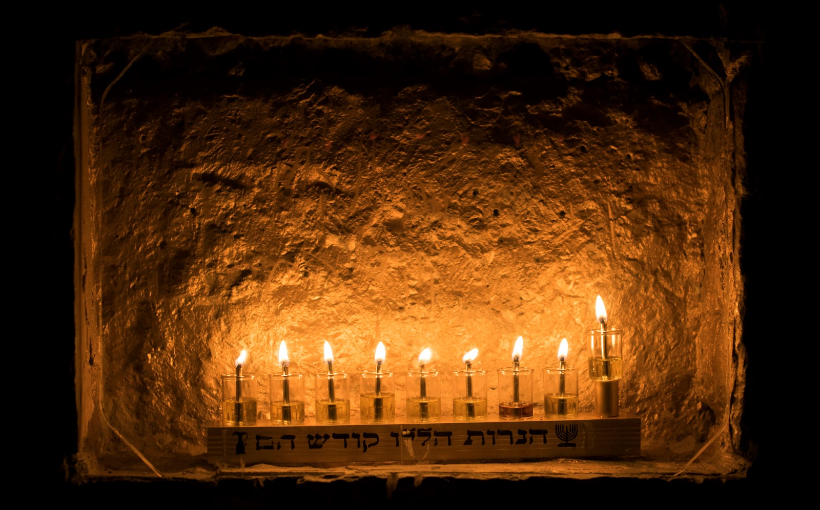 Nine lit candles in glass holders are arranged in a row on a small shelf against a textured, warm-toned wall. Hebrew text is visible on the shelf below the candles, which glow softly in the dim light.