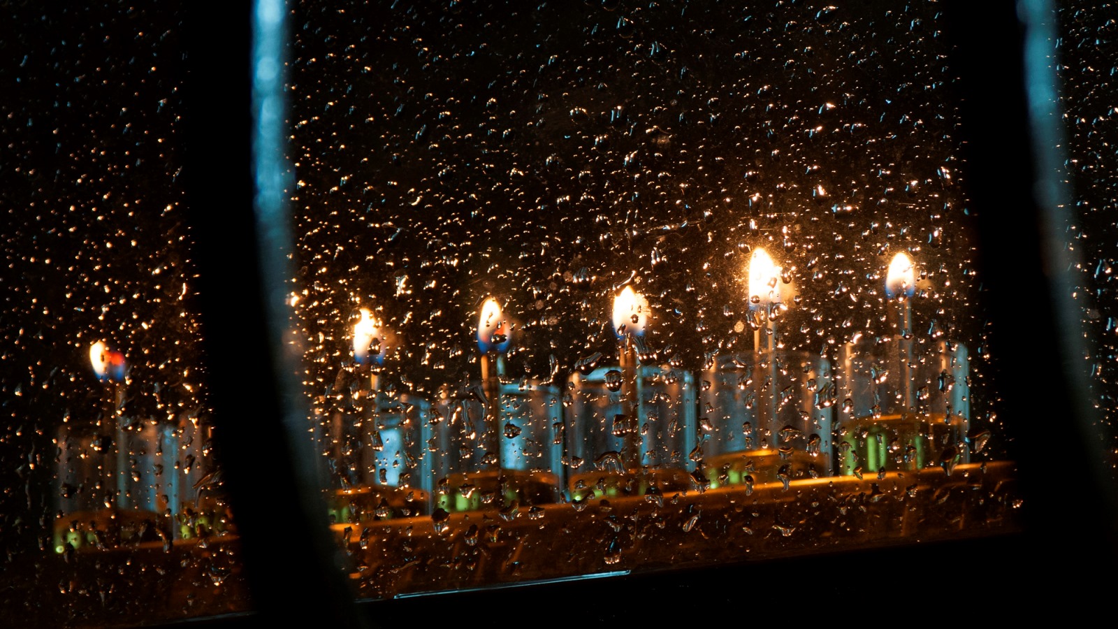 A menorah with candles burning brightly sits behind a rain-speckled window at night, with droplets of water on the glass reflecting the warm candlelight.