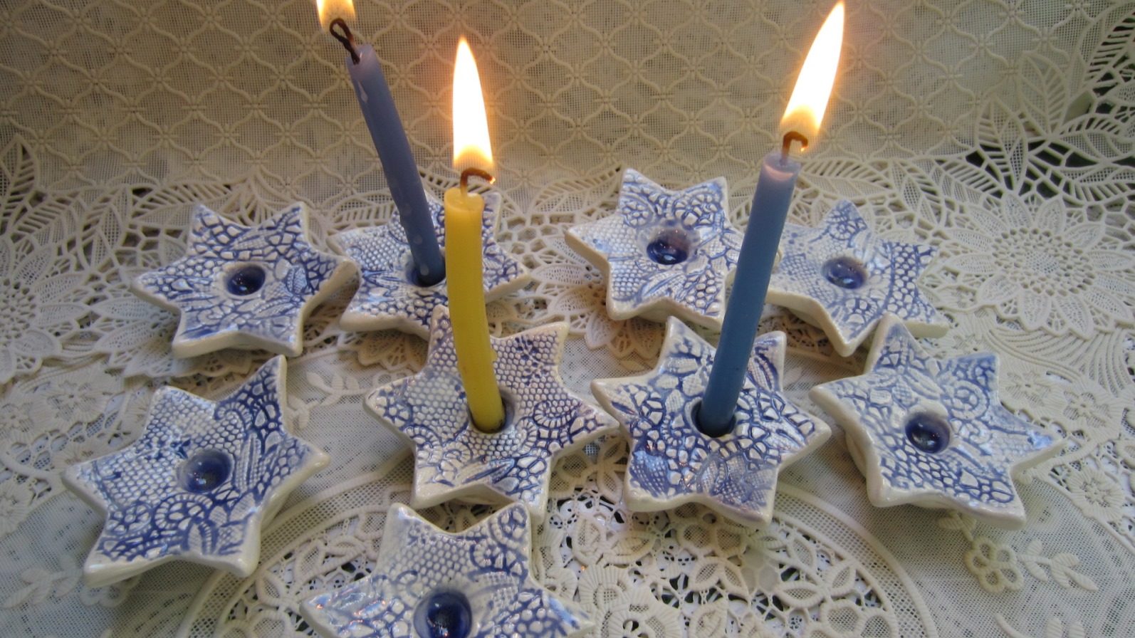 Ceramic Jewish Heirlooms are meant to be passed down across the generations. Photo by Alida Bunder Eight blue-and-white star-shaped ceramic candle holders, each holding a colored candle, are arranged on a lace tablecloth. Two candles are lit, and the holders feature intricate patterns.