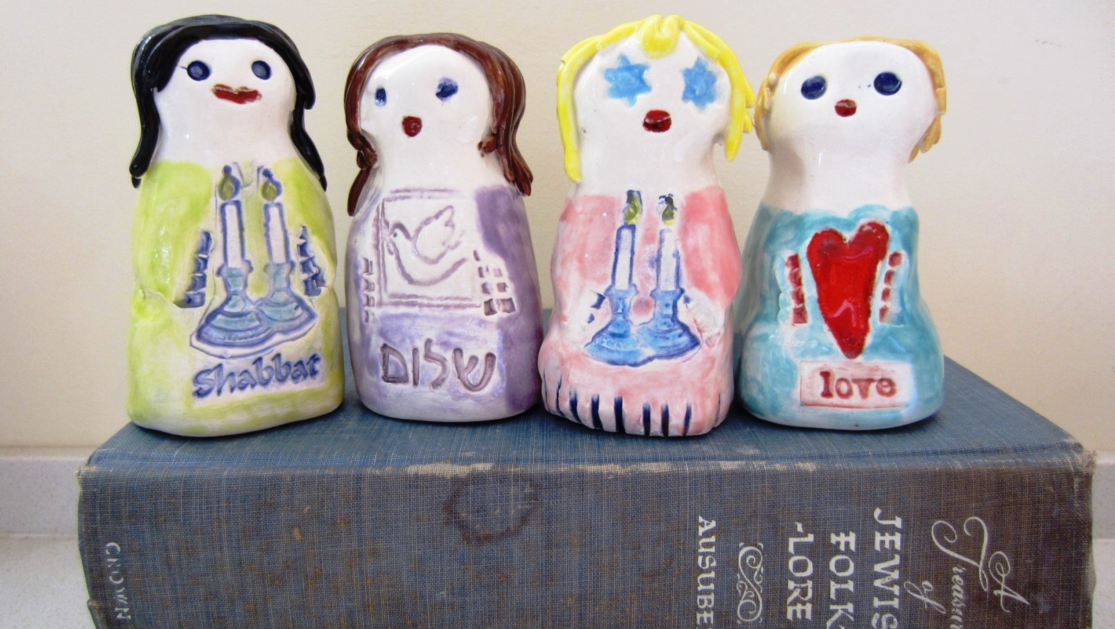 Ceramic Jewish Heirlooms by Alida Bunder. Photo: courtesy Four hand-painted ceramic figurines with candle and heart motifs stand on a vintage book titled Jewish Folklore. Each figure has a unique outfit and hairstyle, with text such as Shabbat, love, and Hebrew writing.