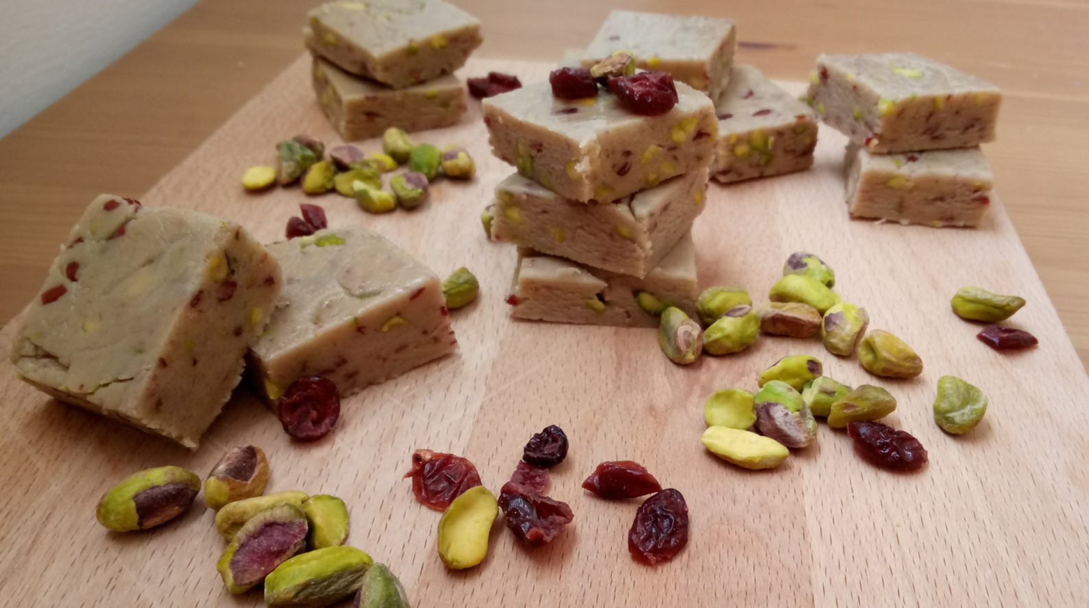 Squares of homemade halva with visible pistachios and dried cranberries stacked and scattered on a wooden board. Loose pistachios and cranberries are also spread around the halva pieces. 