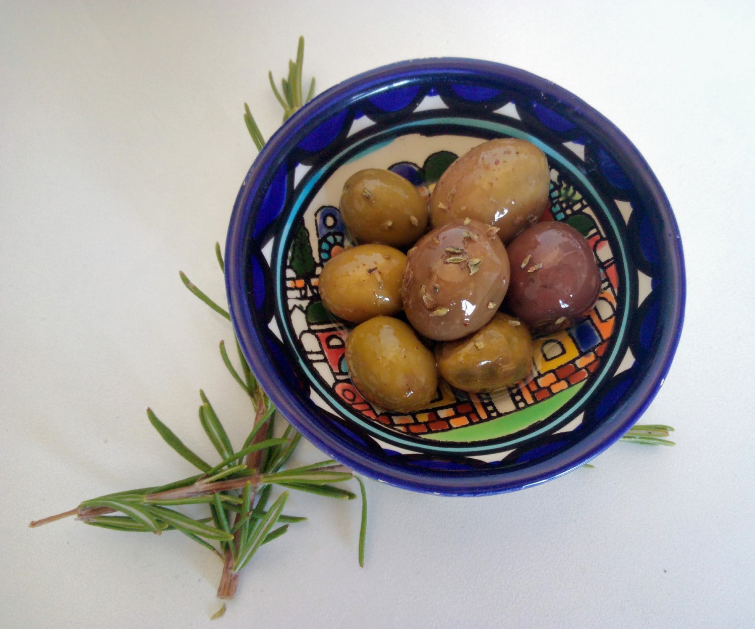 A colorful ceramic bowl filled with assorted marinated olives sits on a white surface, with a few sprigs of fresh rosemary placed beside it. 