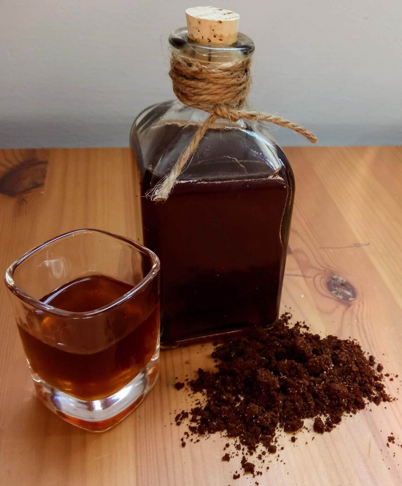 A glass bottle with a cork and twine, filled with Turkish coffee liqueur for the holidays. It sits on a wooden surface next to a shot glass of the same liqueur and a pile of ground Turkish coffee. 