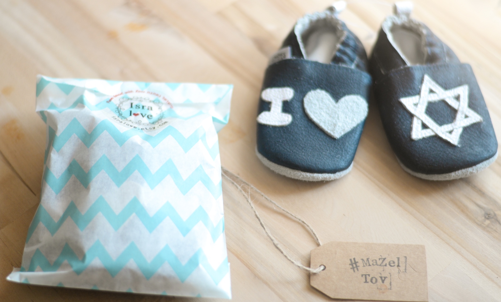 You’ll love Isralove’s baby gifts. Photo: courtesy A pair of baby shoes with a heart and Star of David design sits next to a chevron-patterned gift bag and a tag reading #MazelTov! on a wooden surface.