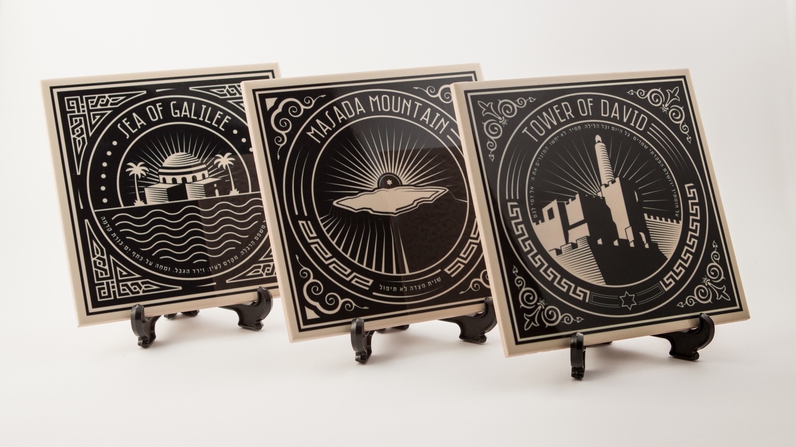 Piece of History Israeli leader figurines. Photo: courtesy Three decorative black-and-white tiles with stylized illustrations on display stands: Sea of Galilee, Masada Mountains, and Tower of David, each labeled with bold text and geometric borders.