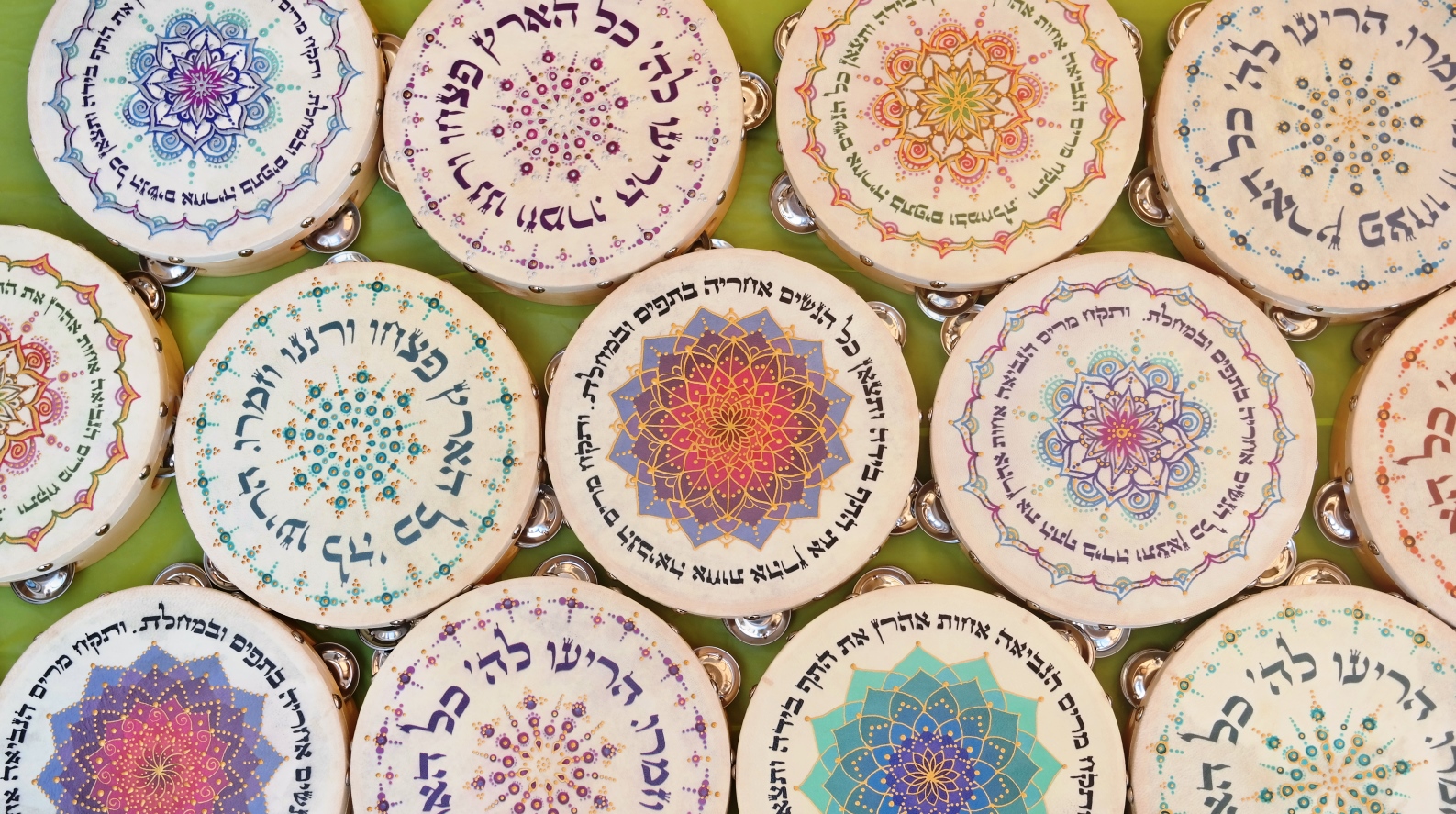 Several decorated tambourines are arranged in rows, each featuring intricate, colorful mandala patterns and Hebrew text on their surfaces against a green background.