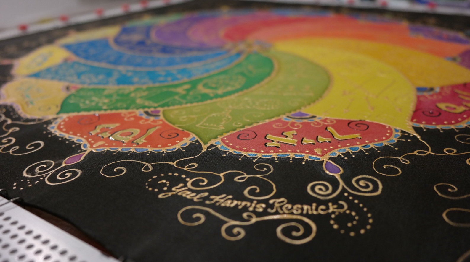 Challah cover by Ibex Art and Design. Photo by Yael Harris Resnick A colorful mandala-like artwork with sections in yellow, green, blue, pink, and purple on a black background, decorated with gold swirls and including the signatures of Yael Harris Resnick at the bottom.
