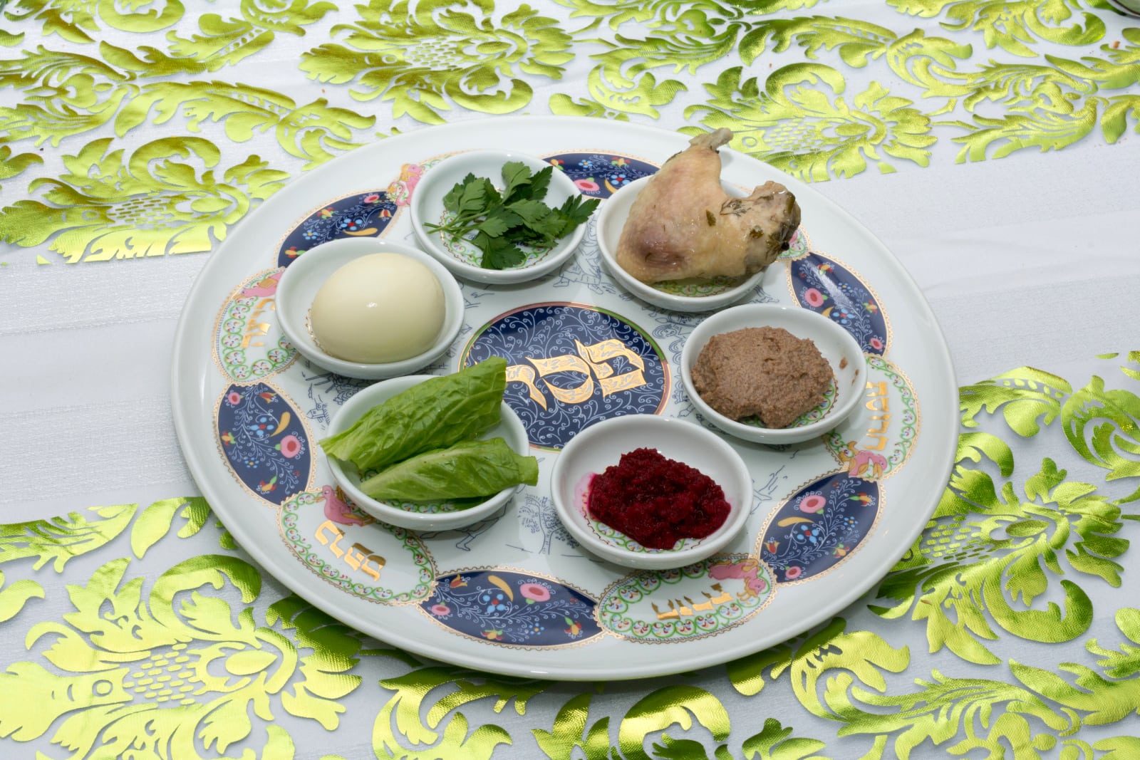 A Passover Seder plate with six traditional symbolic foods arranged in small dishes, placed on a decorative green and white tablecloth.