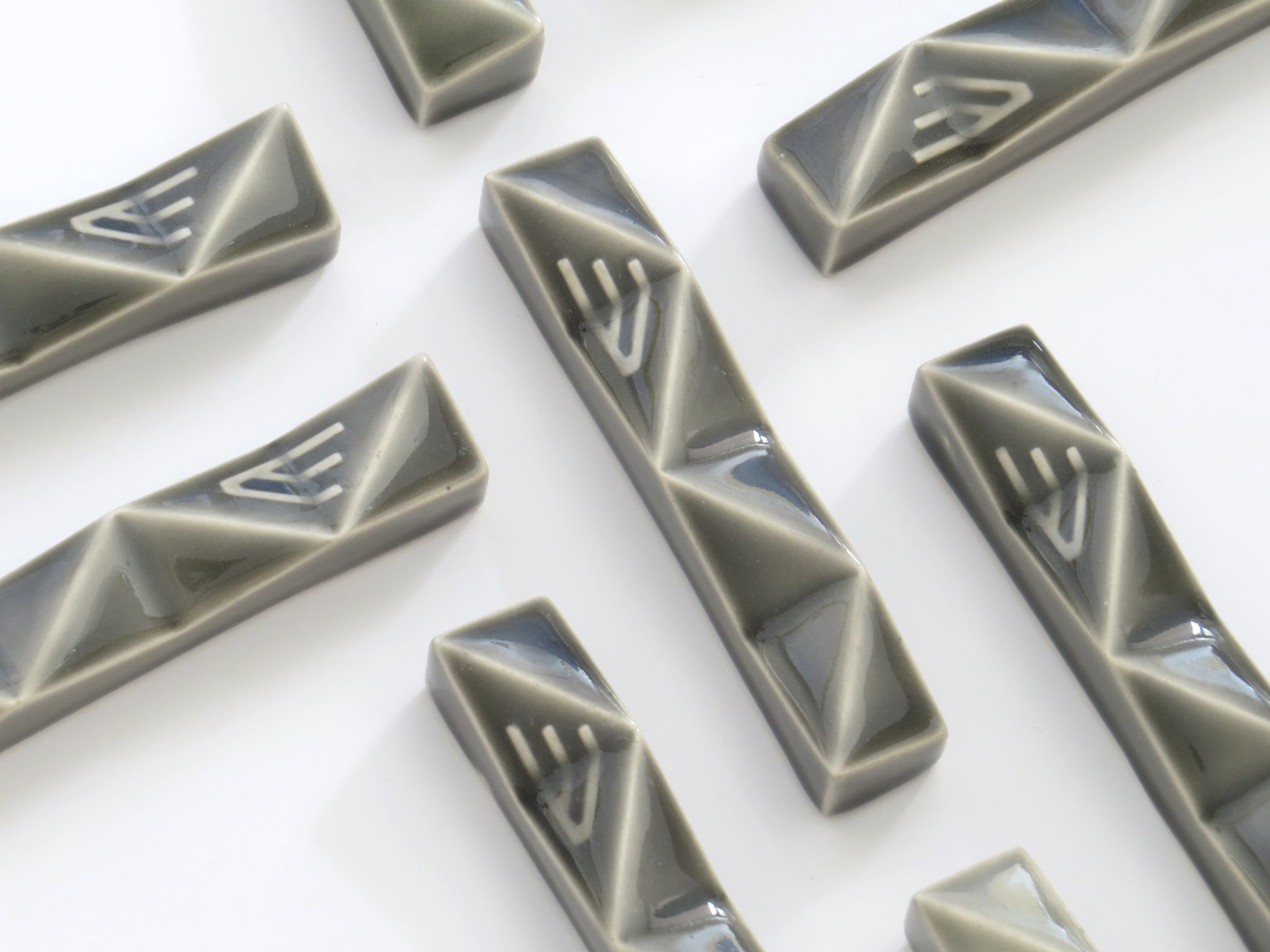 Several glossy, grey ceramic Mezuzot with geometric facets and white abstract line patterns are arranged on a white surface.