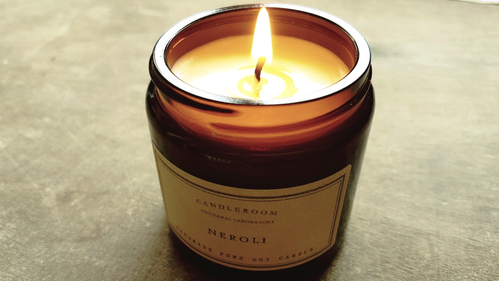 A lit candle in a brown glass jar labeled Candleroom Neroli sits on a smooth, light brown surface. The flame burns brightly, casting a warm glow on the candles creamy wax.