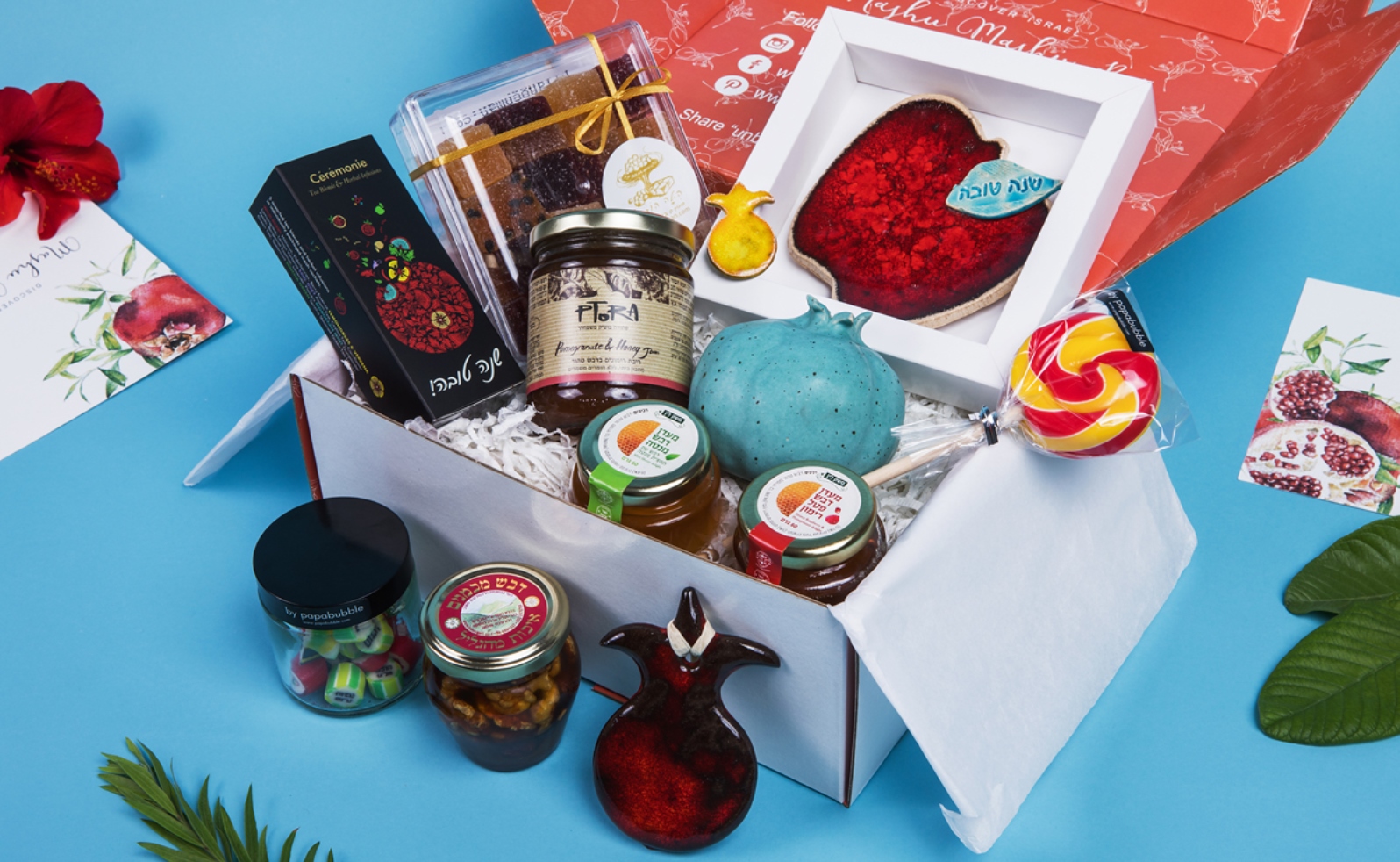 A gift box filled with assorted items, including jars of honey and jam, a lollipop, a blue pomegranate-shaped item, a pomegranate-shaped ceramic, chocolates, and snacks, all displayed on a blue surface with pomegranate-themed cards.