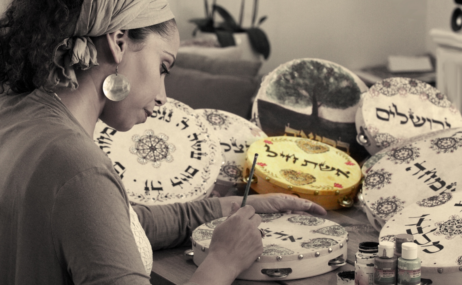 A woman sits at a table, painting intricate designs and Hebrew text on tambourines. Several decorated tambourines surround her, and art supplies are visible nearby.